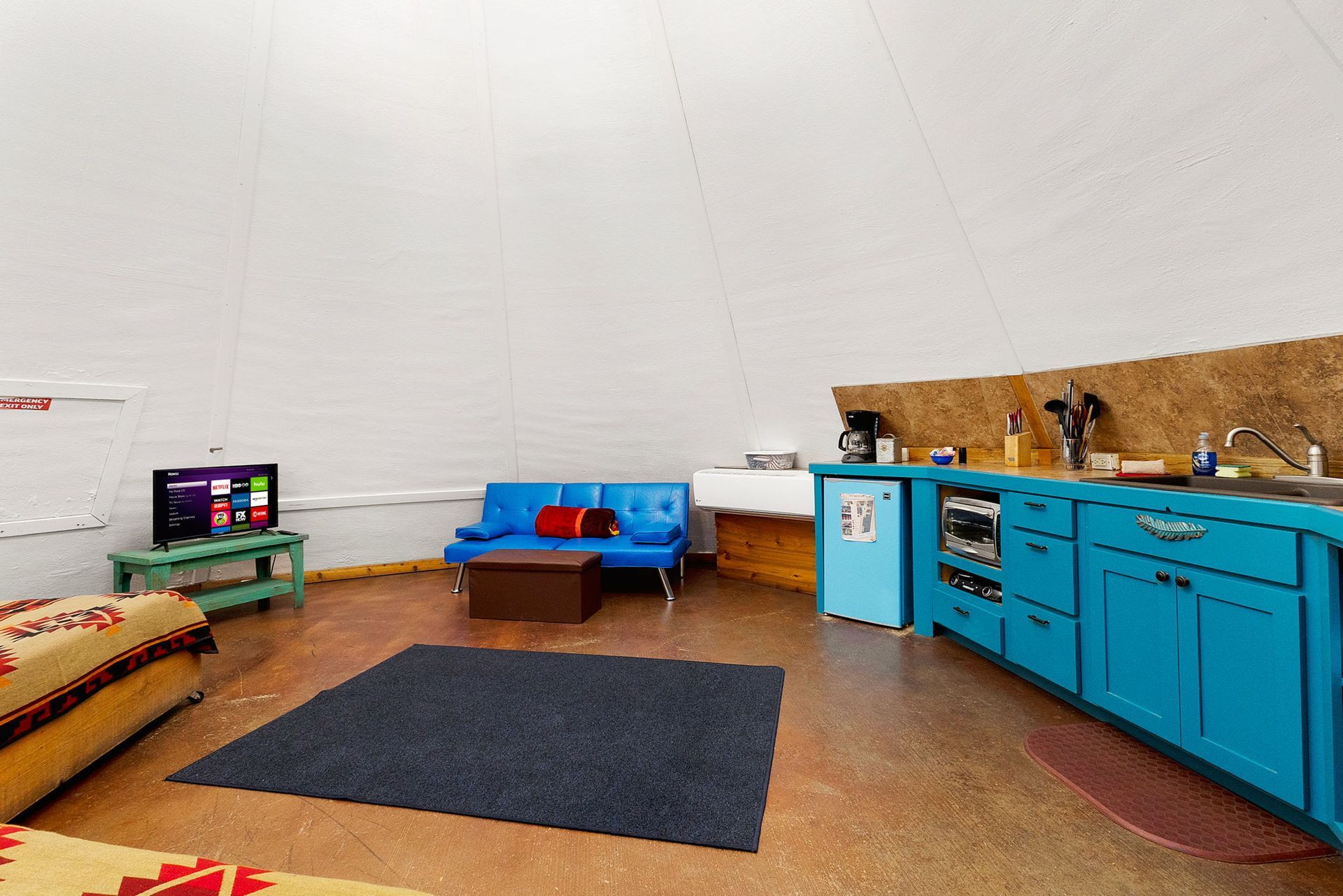 Interior of a yurt with teal kitchen, blue sofa, TV, and dark blue rug.
