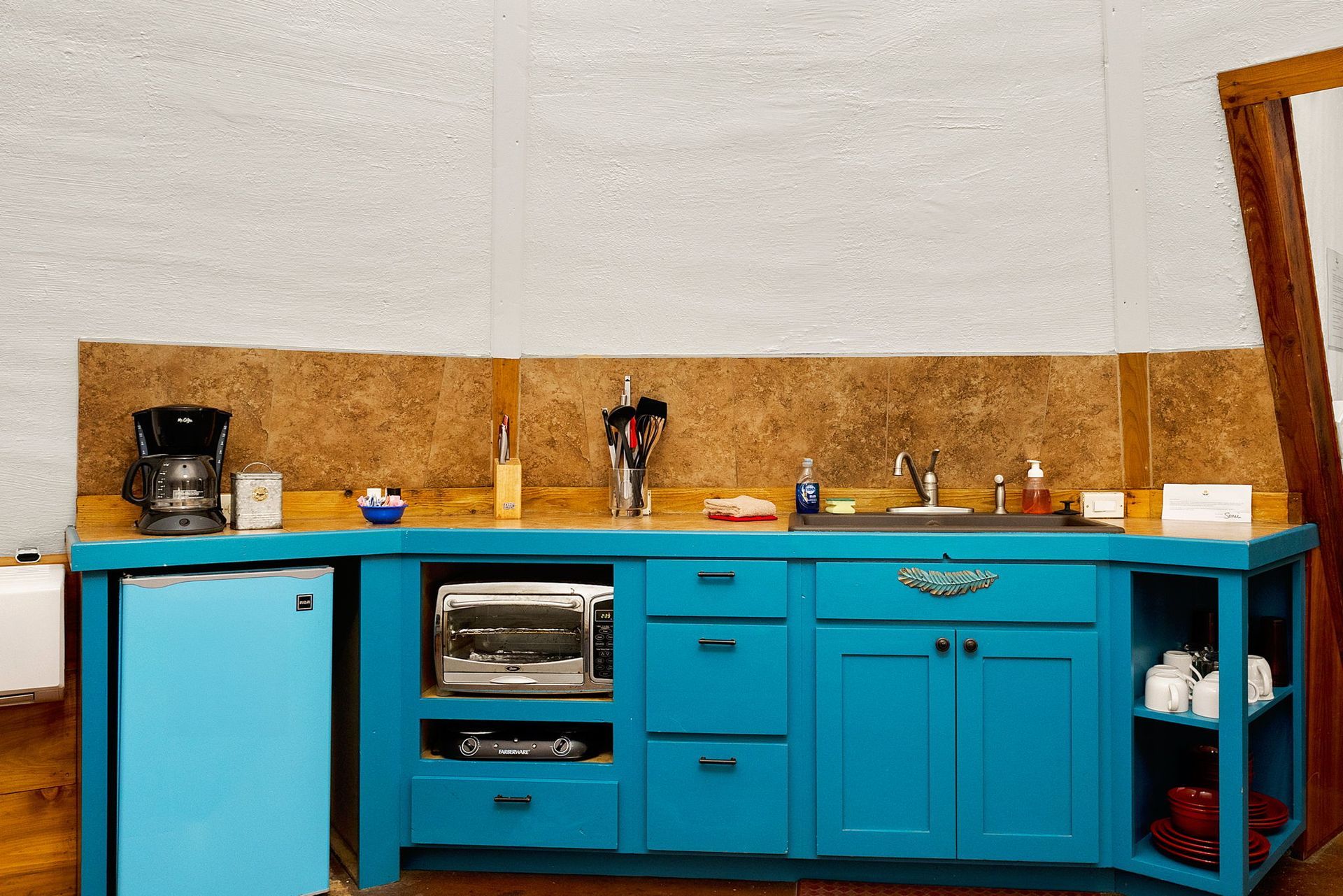 Blue kitchen counter with a refrigerator, toaster oven, and sink against a beige wall.