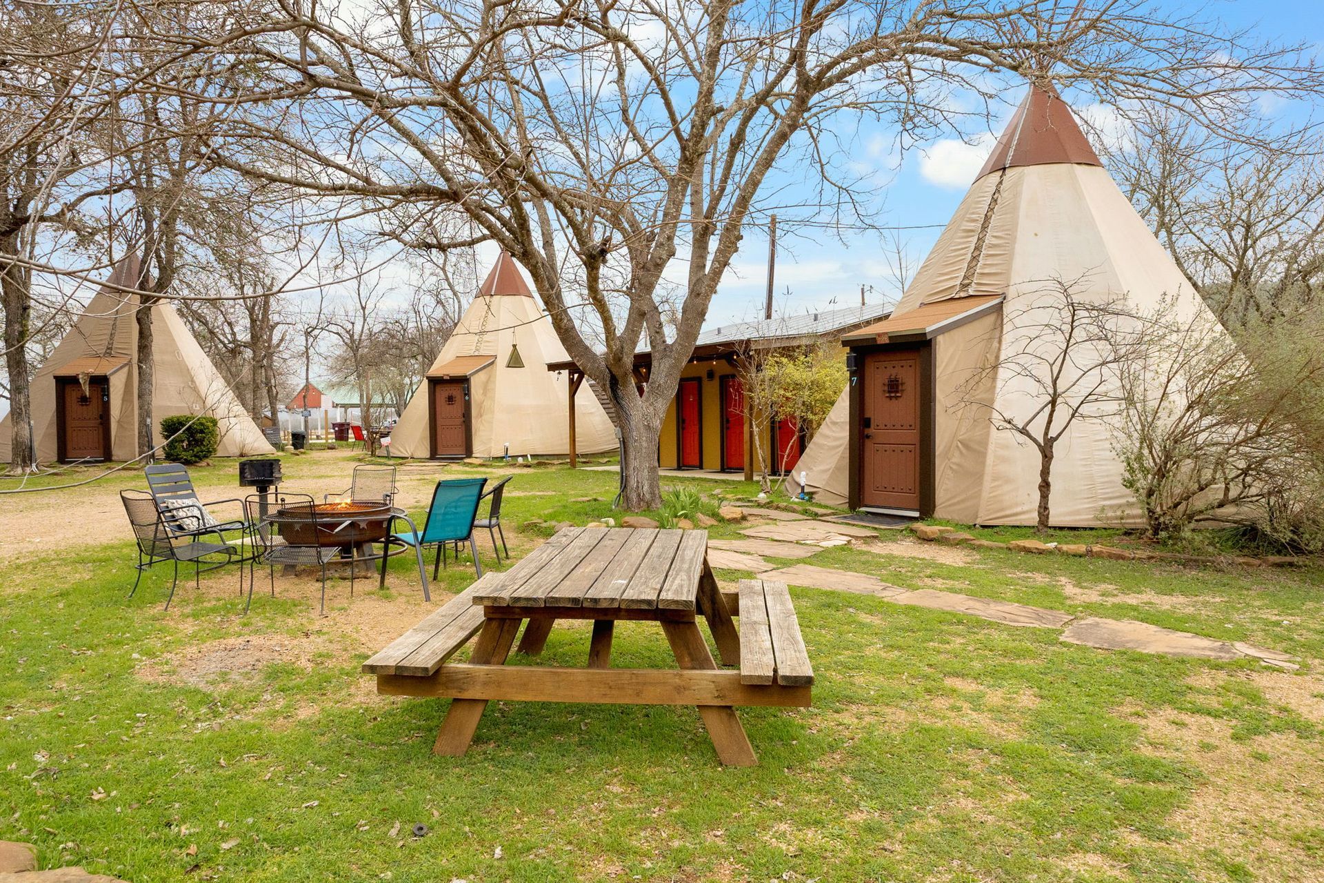 Teepees, picnic table, and fire pit in a grassy yard. Brown doors and tan structures with a clear sky.
