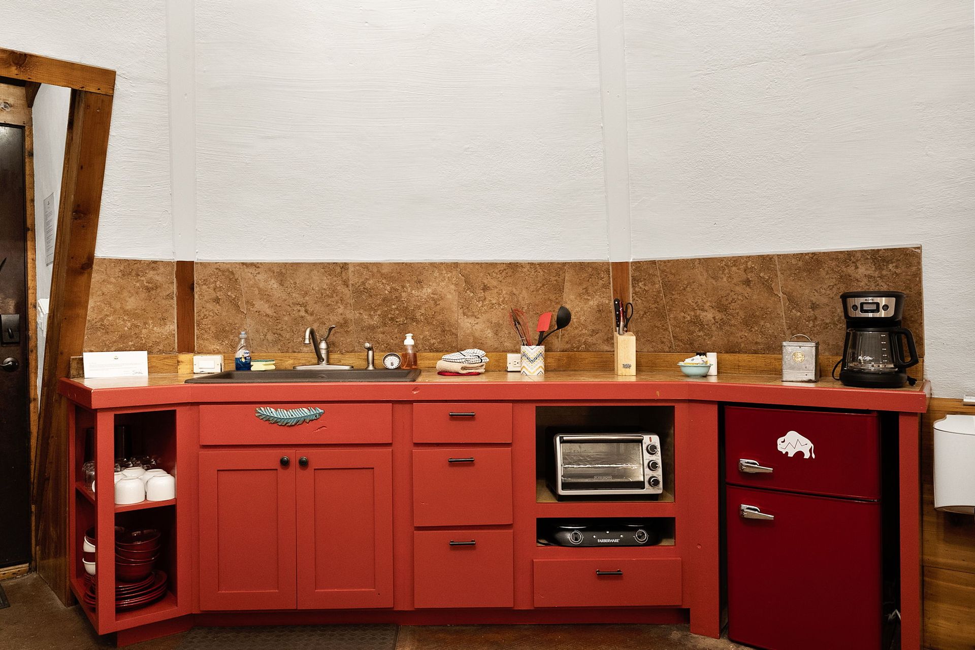 Red kitchen cabinets with sink, microwave, mini-fridge, and coffee maker against a textured backsplash.