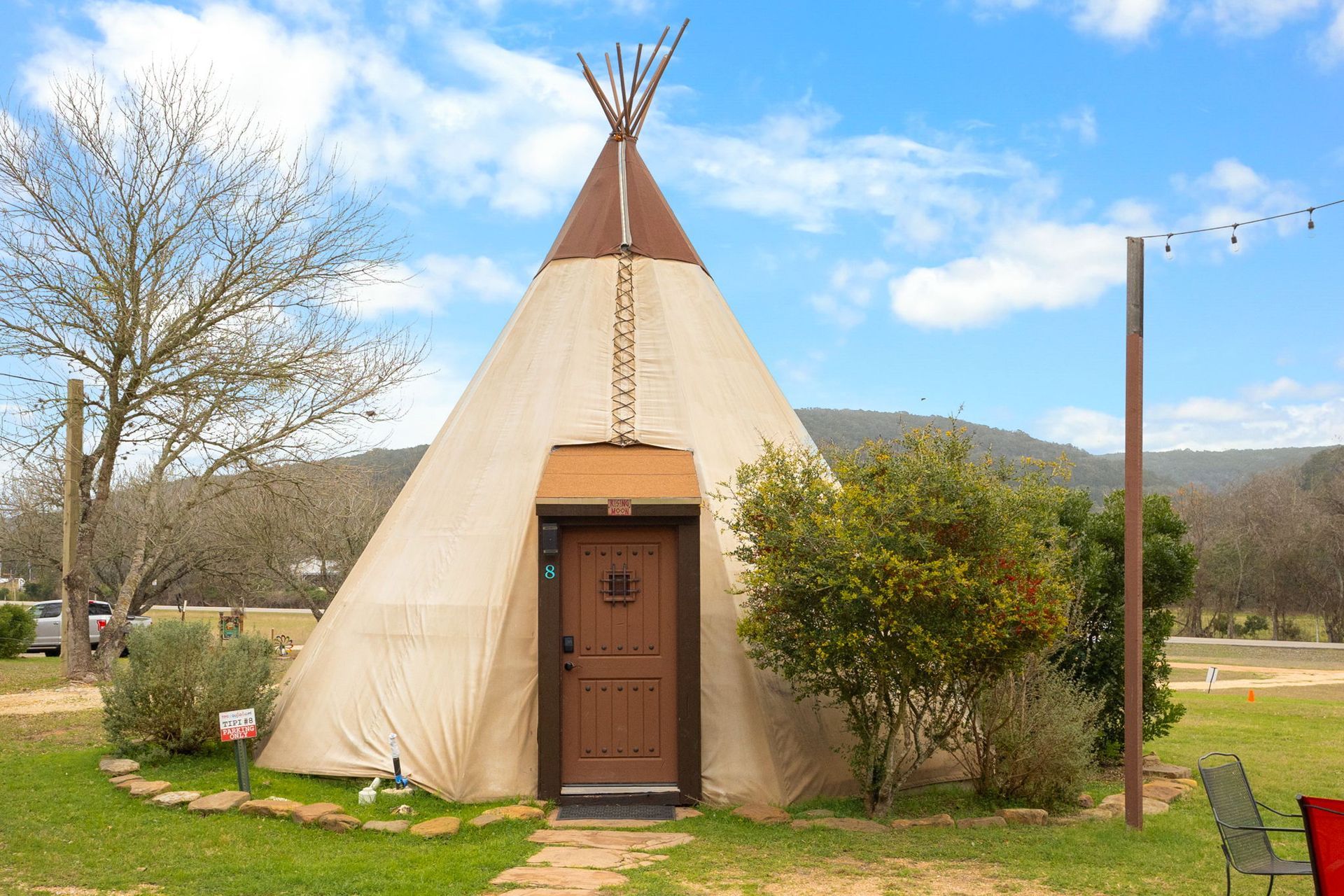 Teepee with a brown door, on a grassy plot. Hills and a tree are in the background. Cloudy sky.