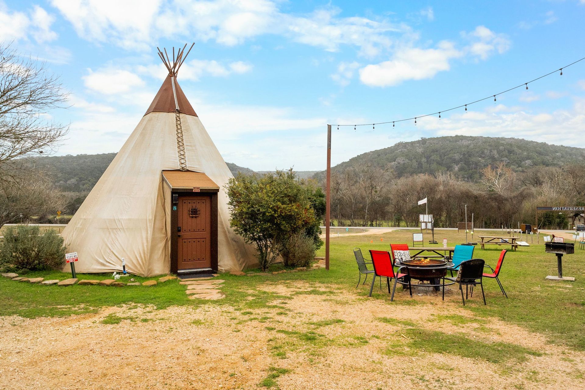 Teepee with a wooden door, outdoor seating, and a grill on a grassy area near hills under a blue sky.