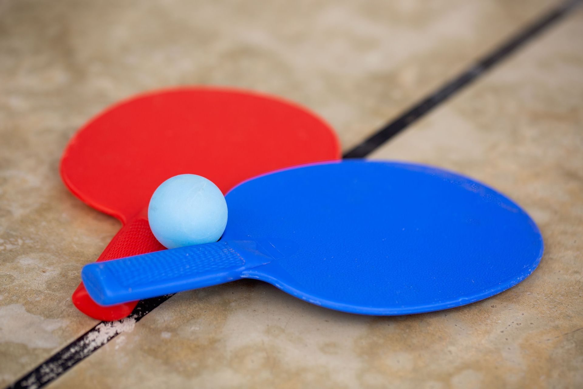 Red and blue ping pong paddles with a light blue ball resting on the blue paddle, set on a brown surface.