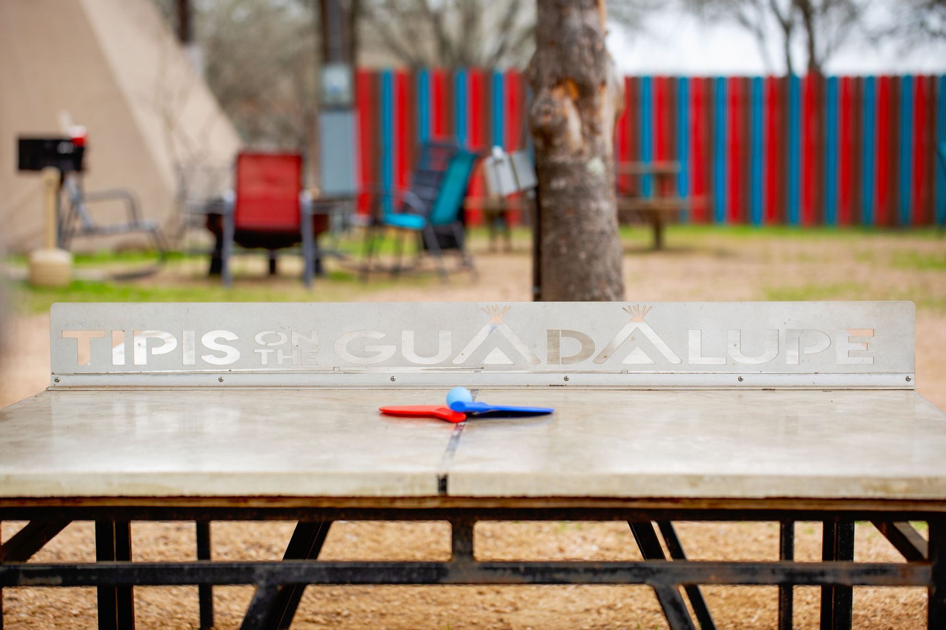 Ping pong table with paddles and Guadalupe signage, outdoor setting with colorful fence.