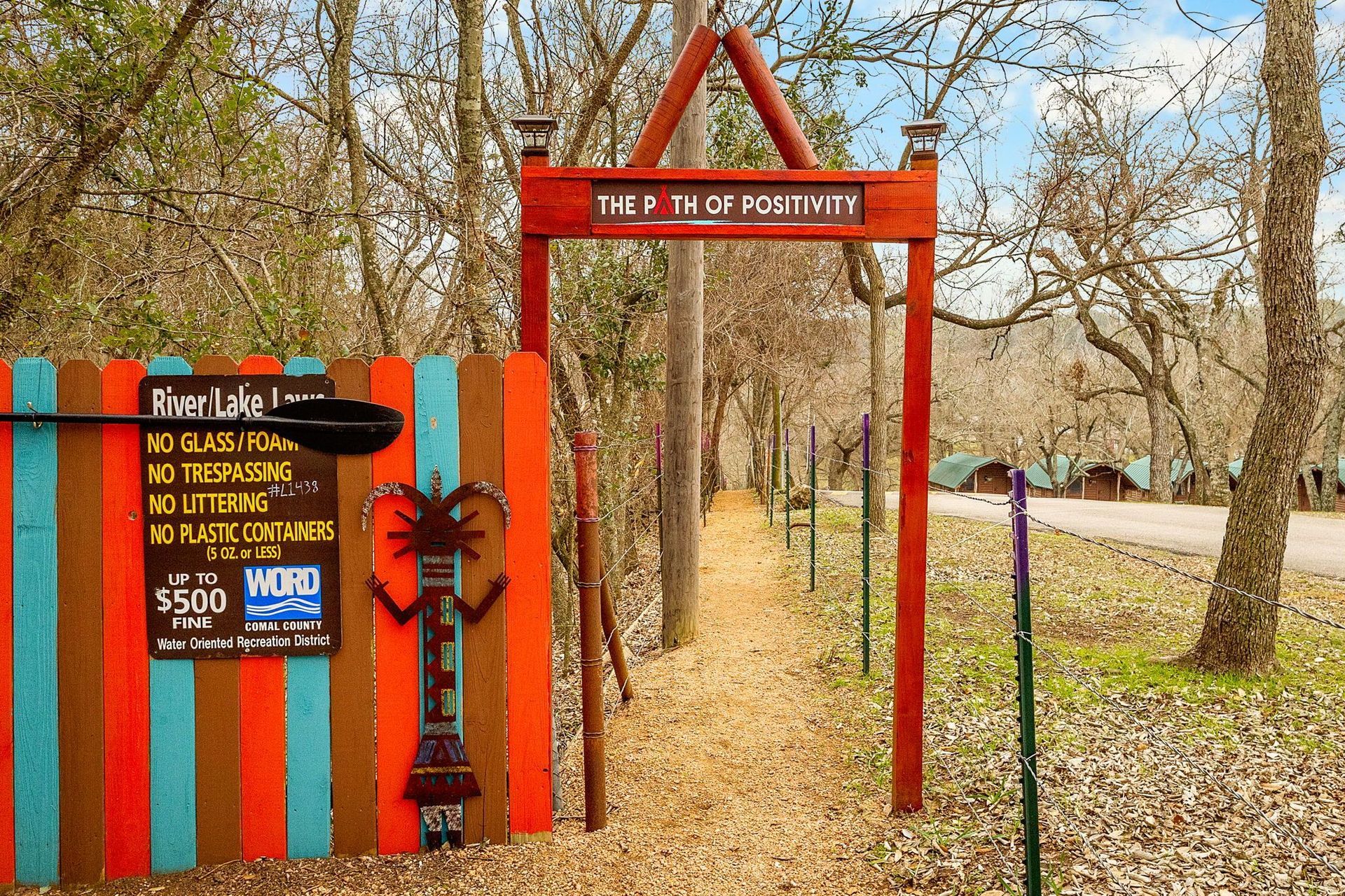 Gate with colorful planks, sign, and pathway leading to cabins, possibly a campsite.