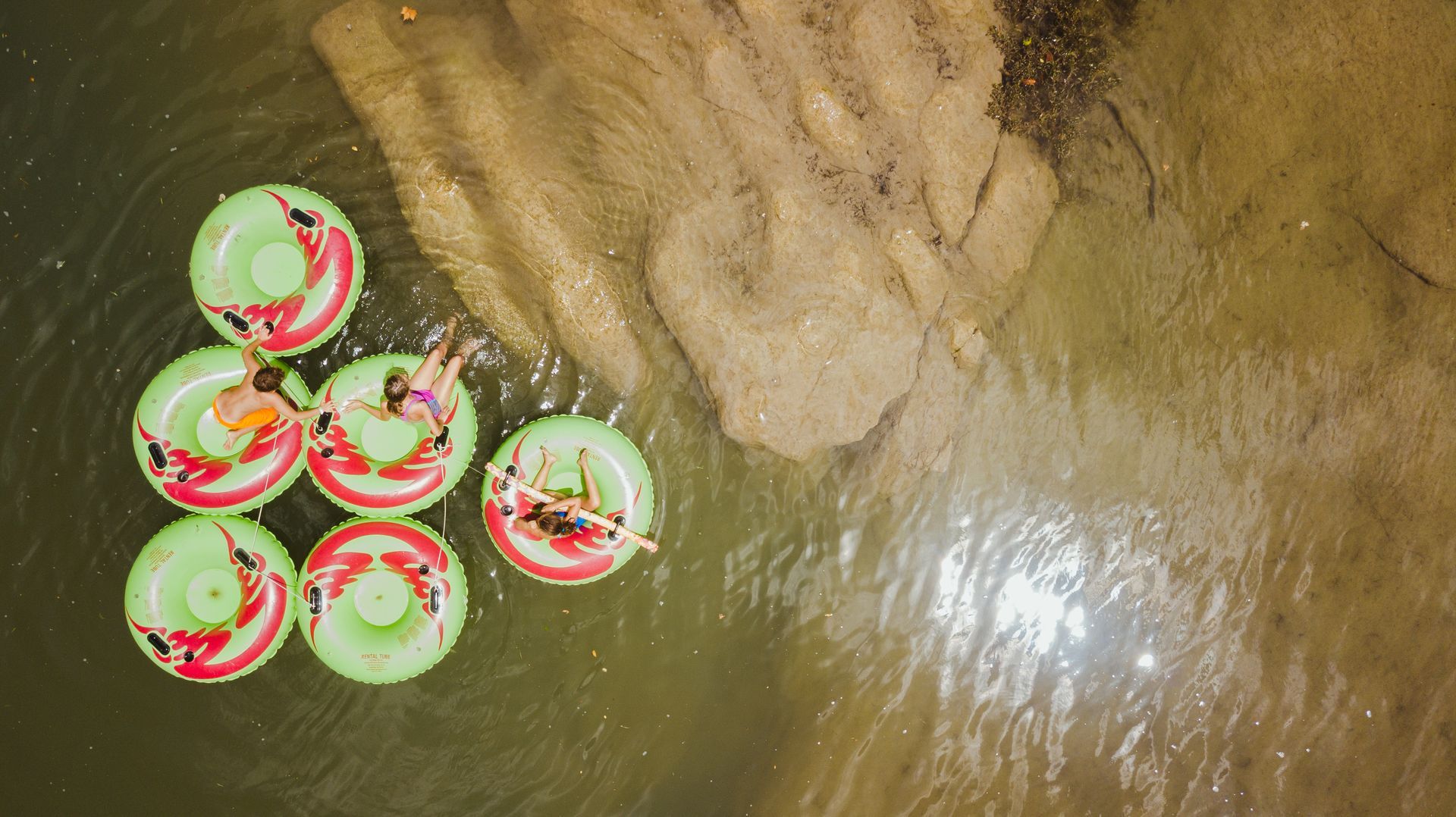 People floating on green and red inner tubes in a river, viewed from above.