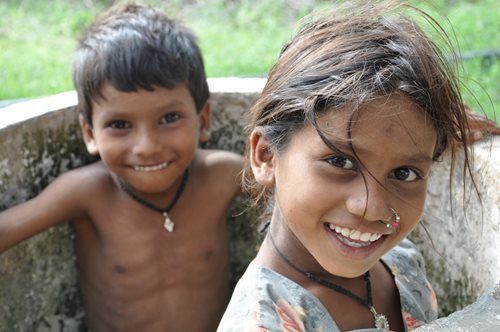 Two smiling children wearing necklaces, near a weathered concrete structure.