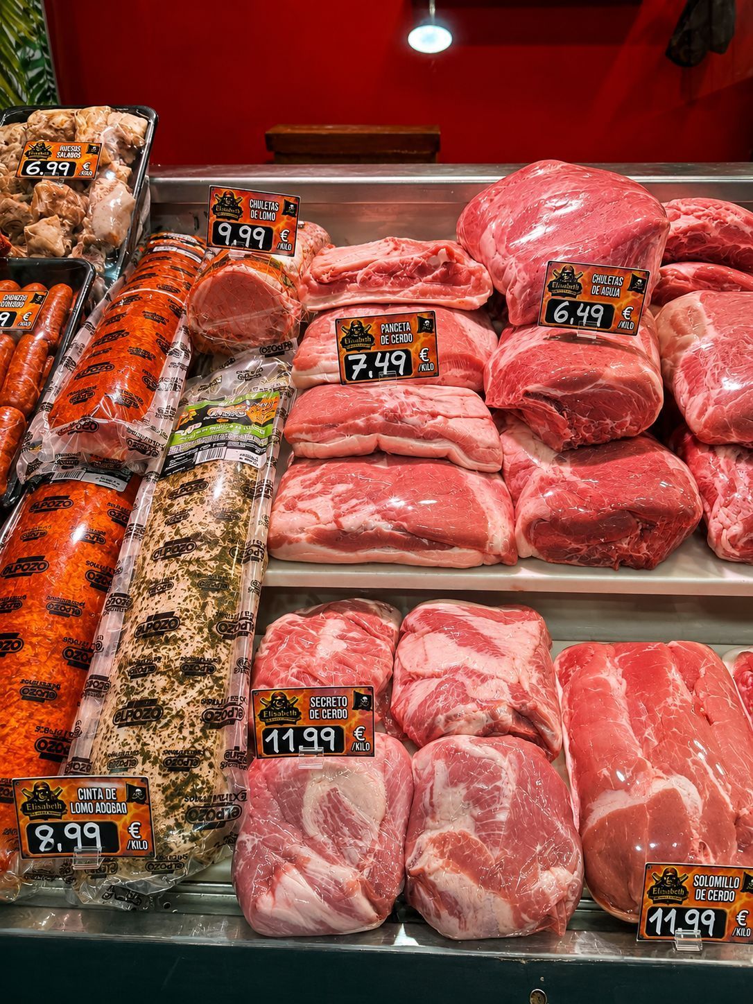 Mostrador de carnicería con bandejas de cortes de carne cruda y etiquetas de precio bajo iluminación roja.