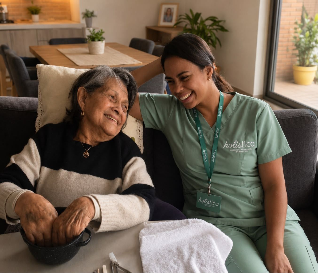 Dos mujeres sonriendo en un sofá en una cálida sala de estar, una con uniforme verde junto a una anciana con una manta.