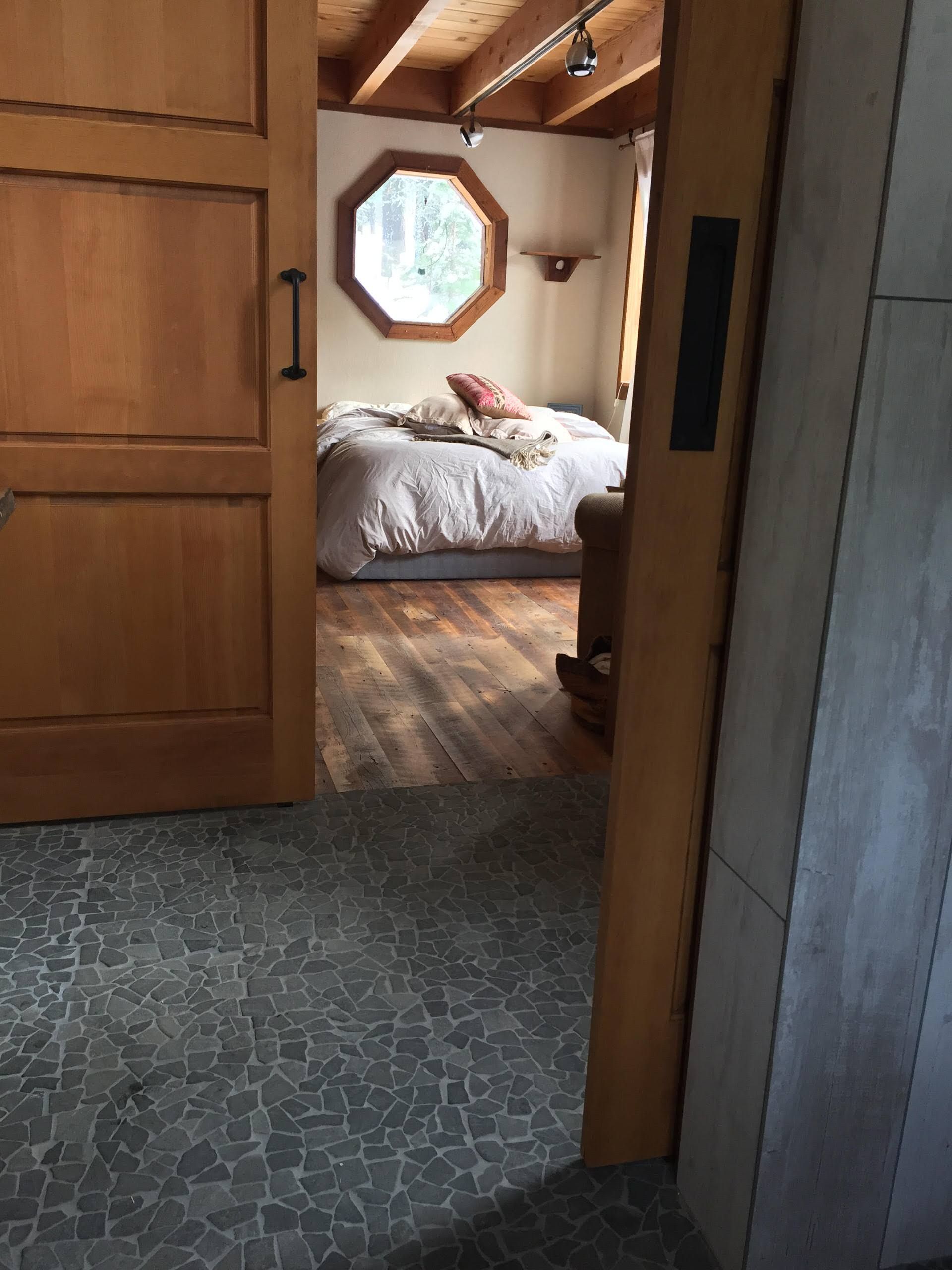 Bedroom interior with wooden door, octagonal window, bed, and visible ceiling beams. Dark gray tiled floor.