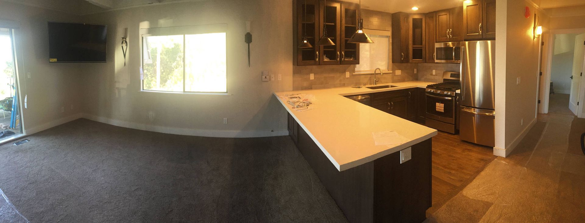 A kitchen with dark cabinets, stainless steel appliances, and a white countertop. Adjacent is a living area with a tv.