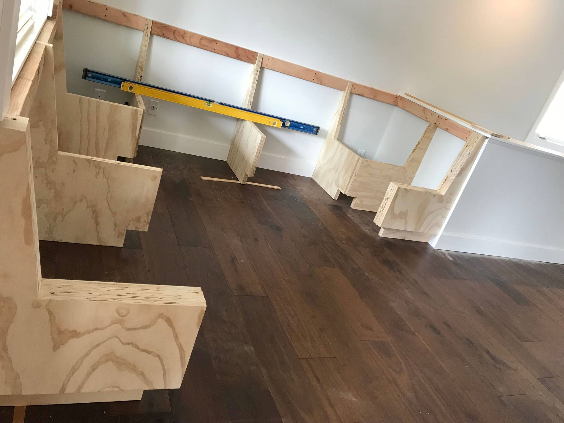 Wooden bench frame being built in a room, angled against a white wall and window. A level is used.