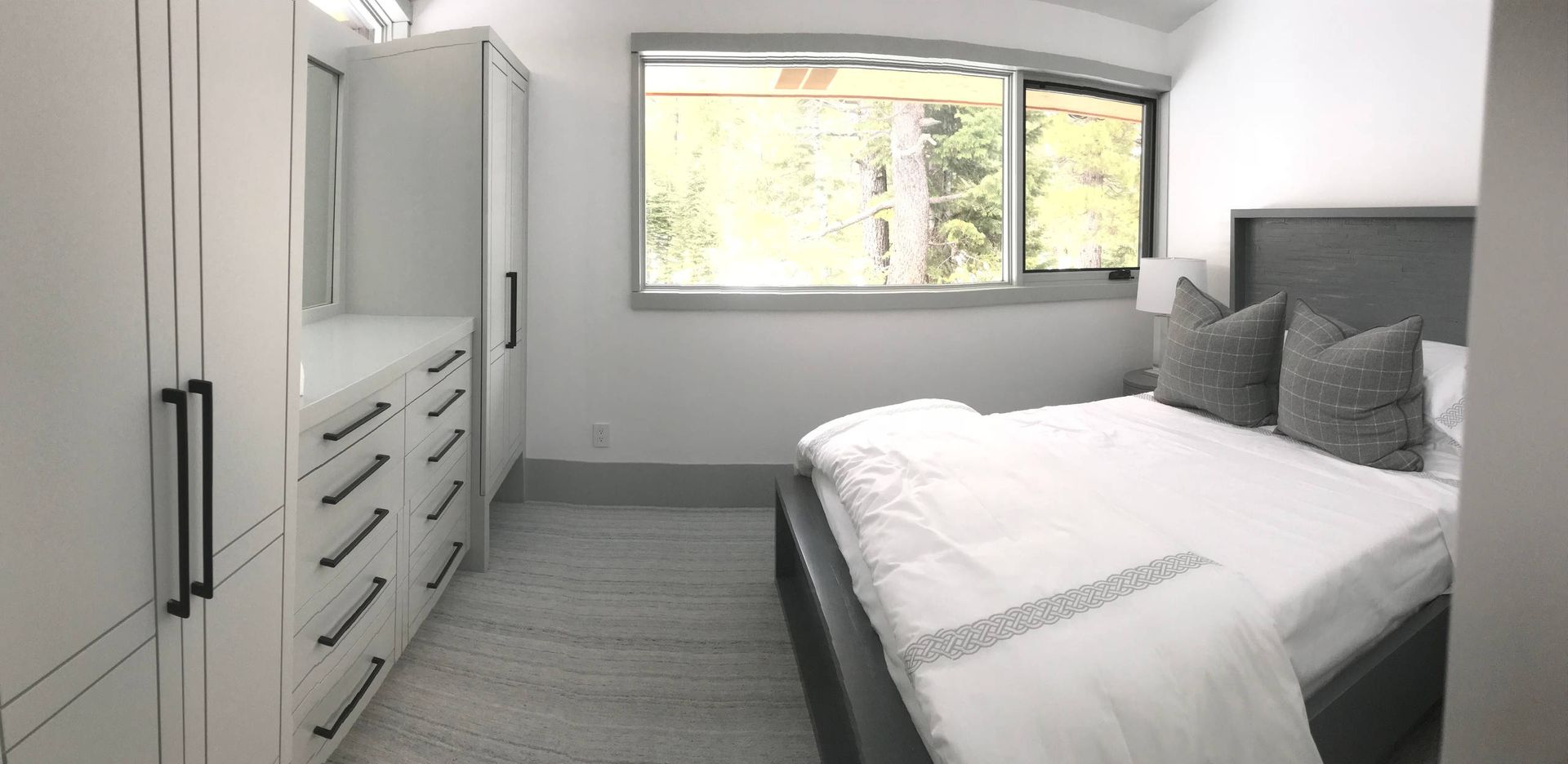 Bedroom with white wardrobes, drawers, bed, window with trees outside.