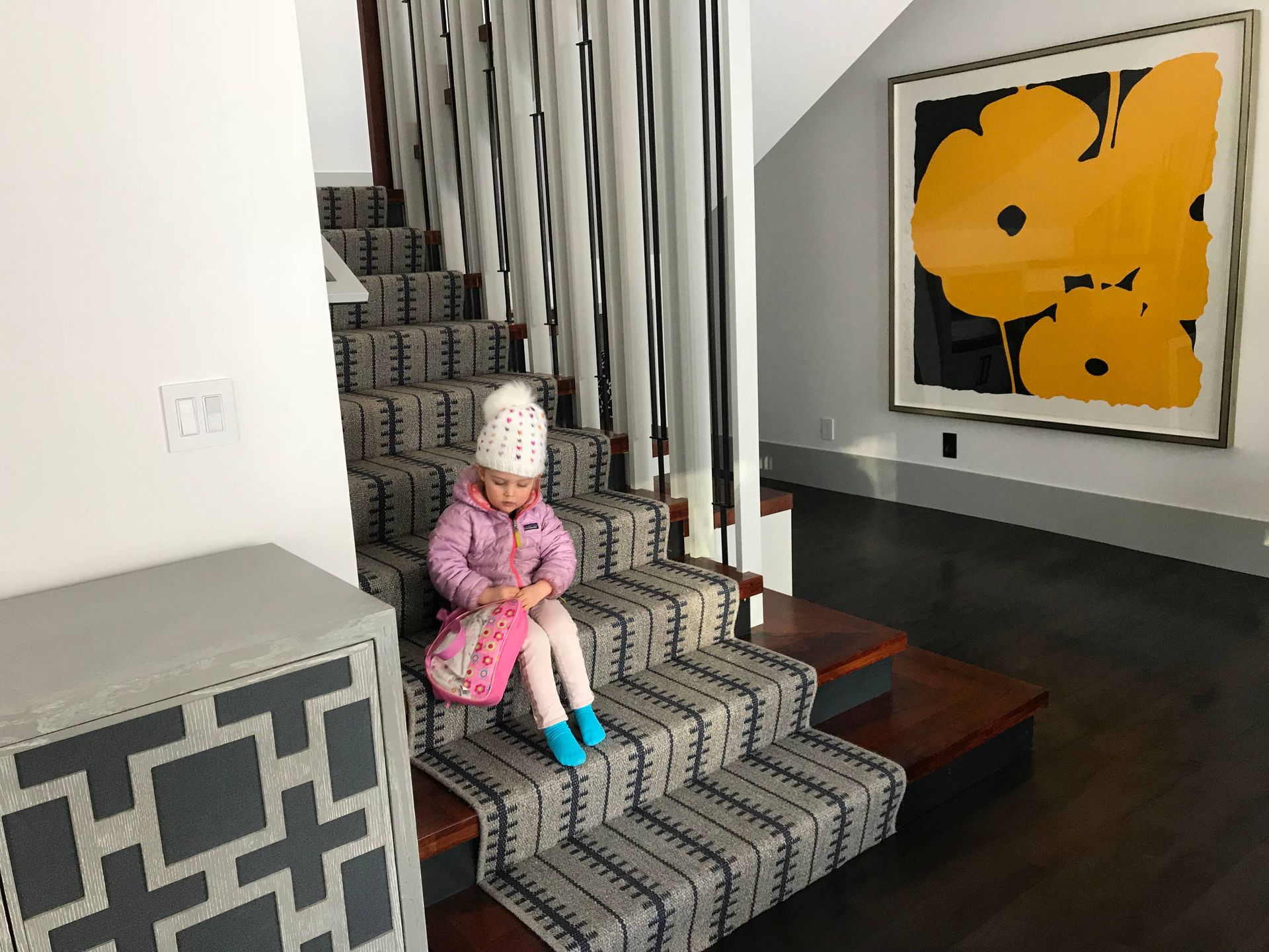 Child in pink jacket and hat sits on staircase with patterned runner, framed artwork with yellow flowers.