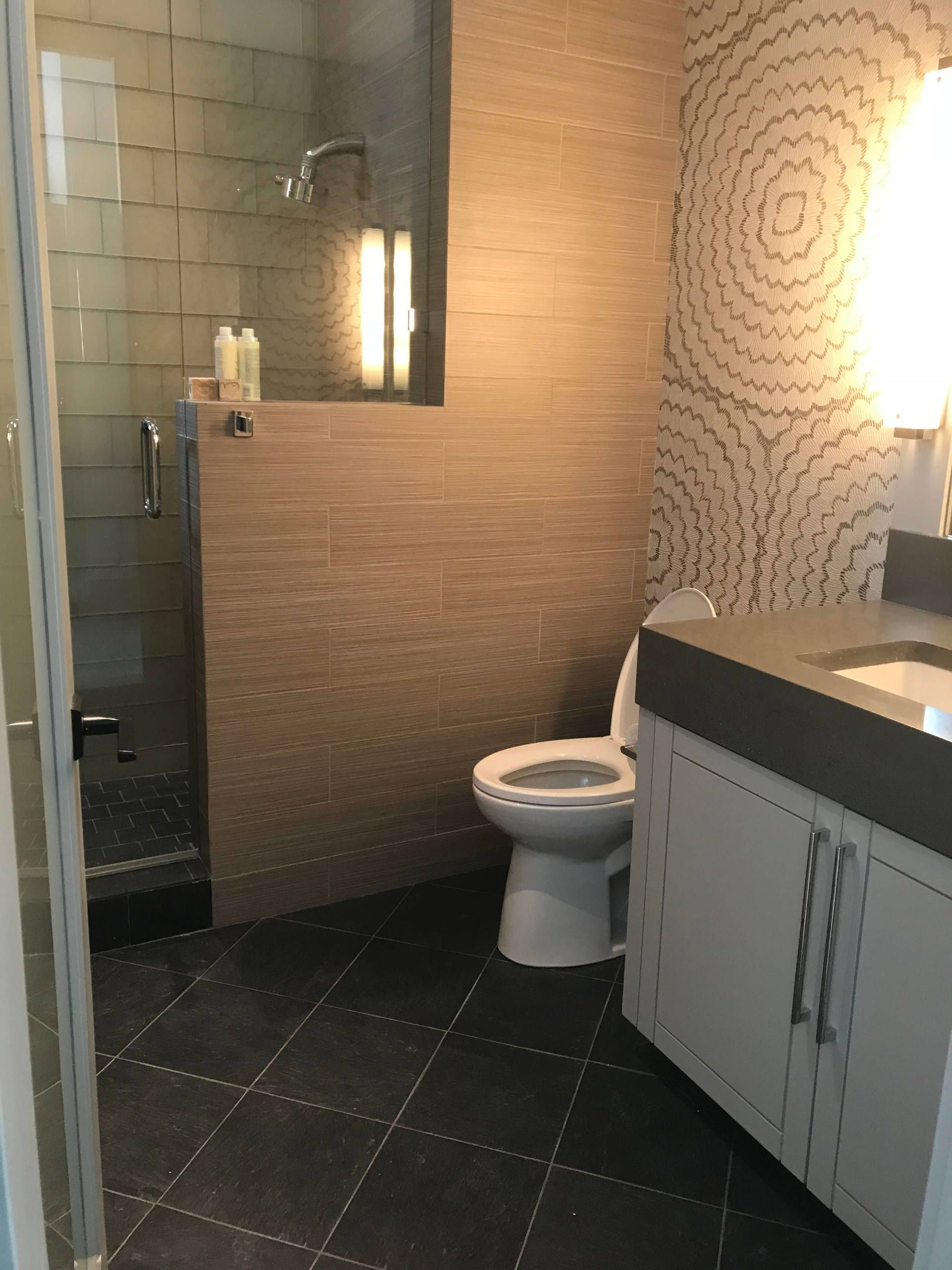 Modern bathroom with shower, toilet, and vanity. Gray tile floor, light walls, and decorative patterned wallpaper.