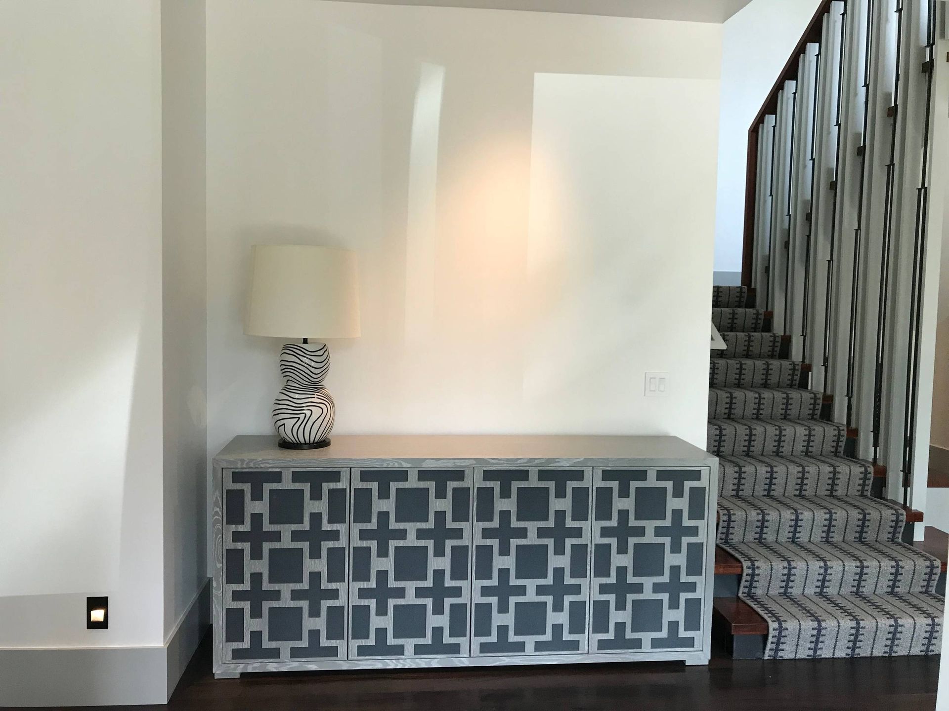 Gray patterned cabinet with lamp, next to staircase. White walls, dark wood floor.