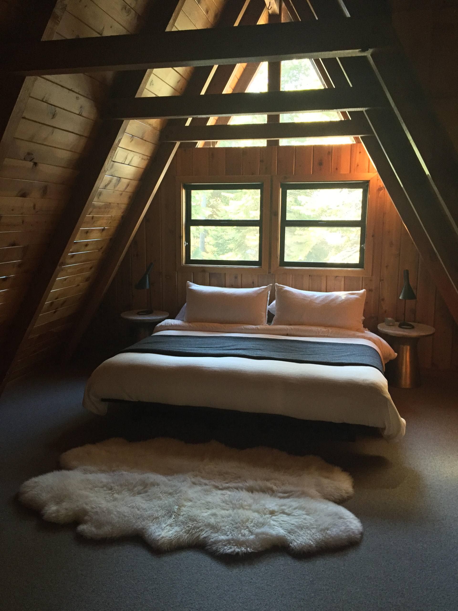 Bedroom in an A-frame cabin. Bed with white linens, grey throw, two windows, sheepskin rug.