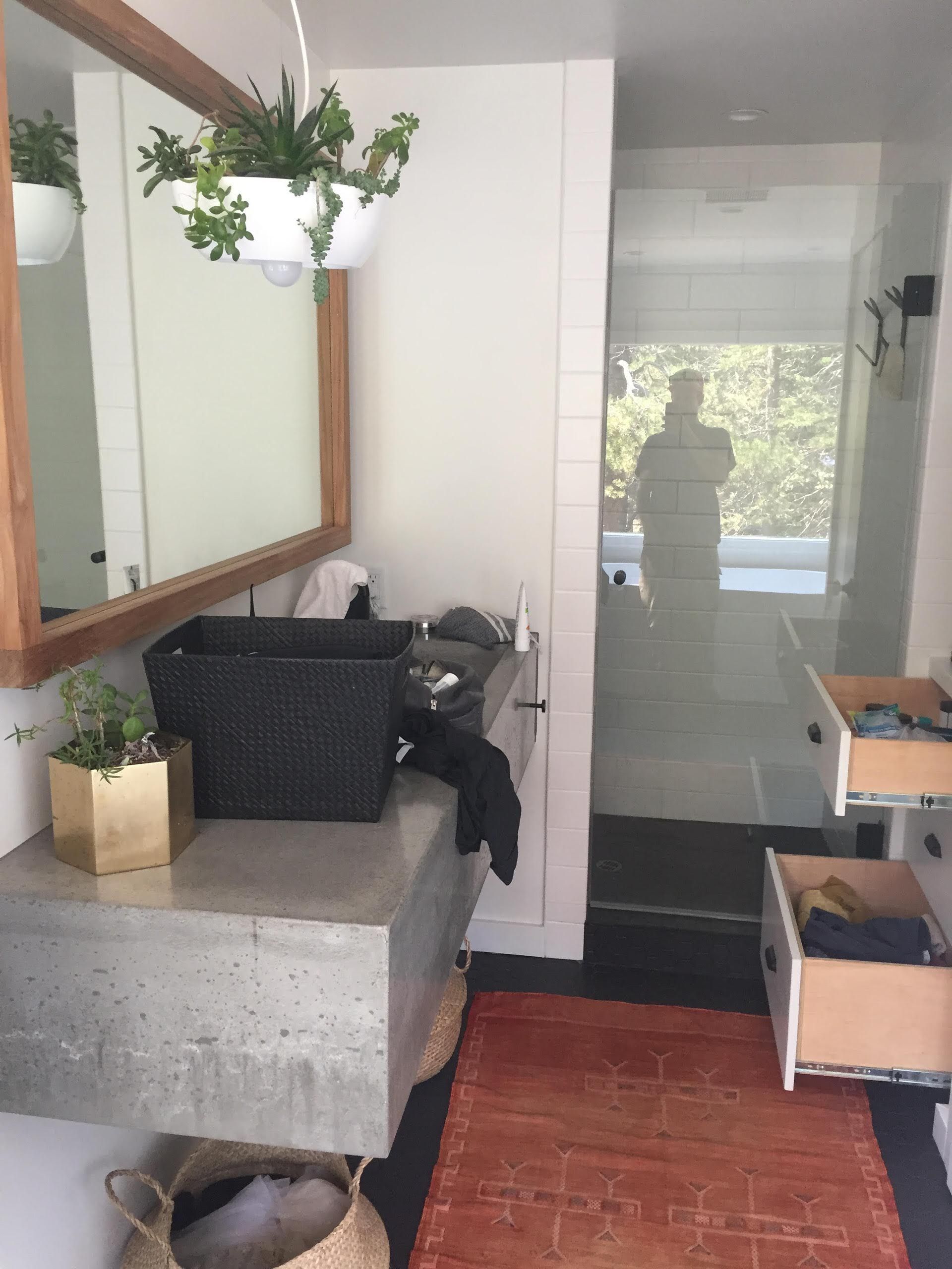 Bathroom with concrete sink, plants, mirror, and open drawers; a person is reflected in a glass door.