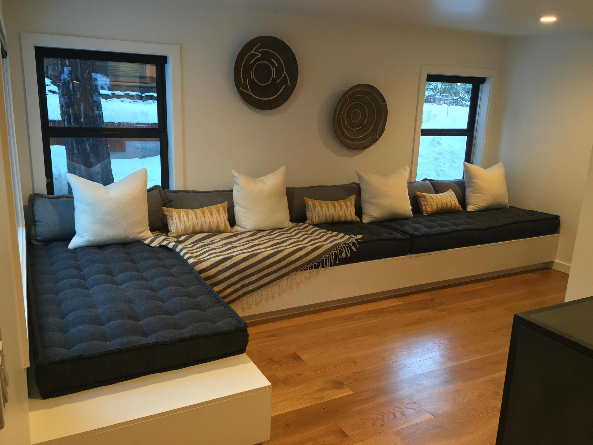 Built-in gray sofa with white pillows and a striped blanket, next to windows with wood flooring.