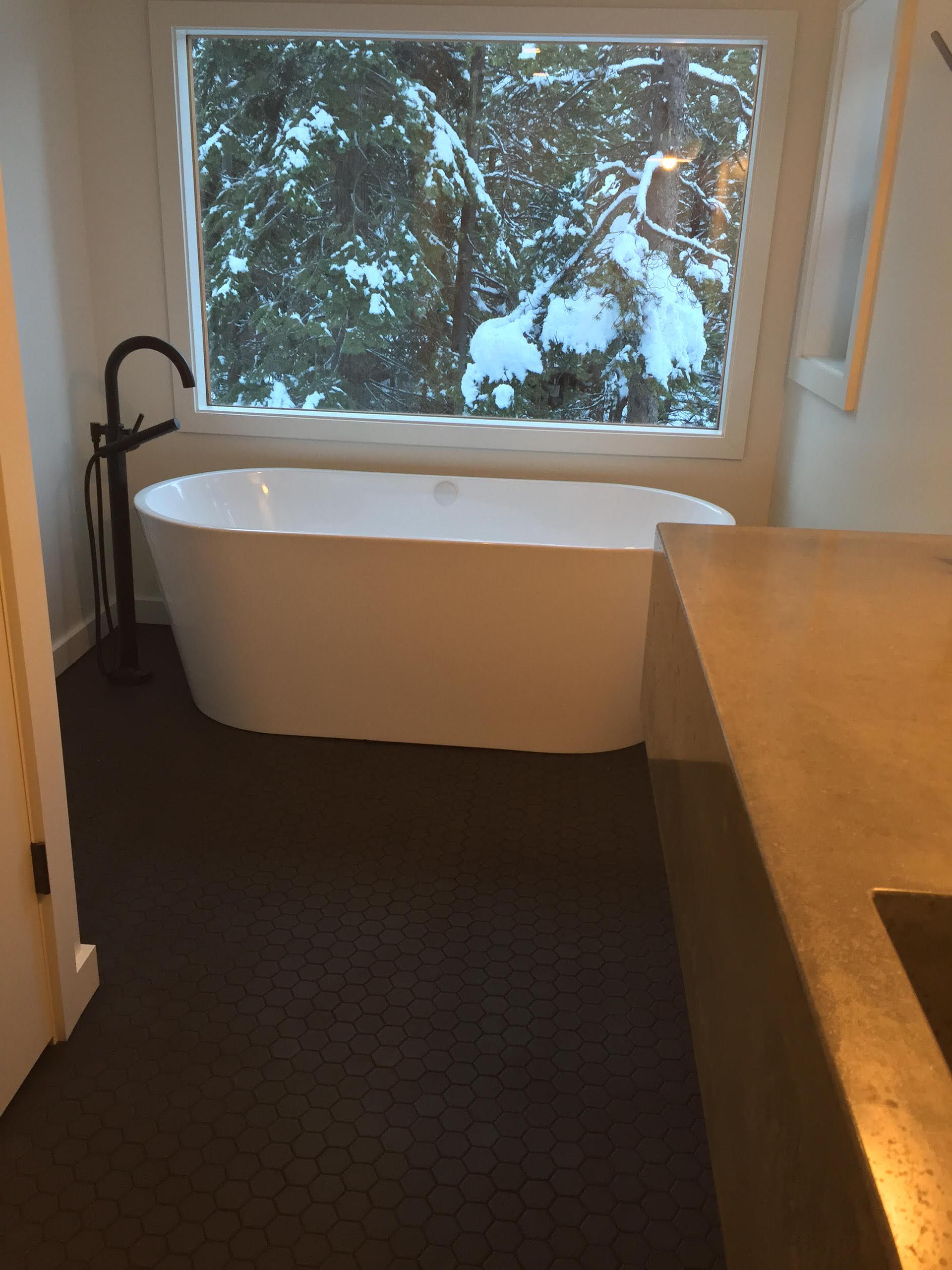Freestanding white bathtub in bathroom, large window with snowy trees outside. Dark floor, black faucet.