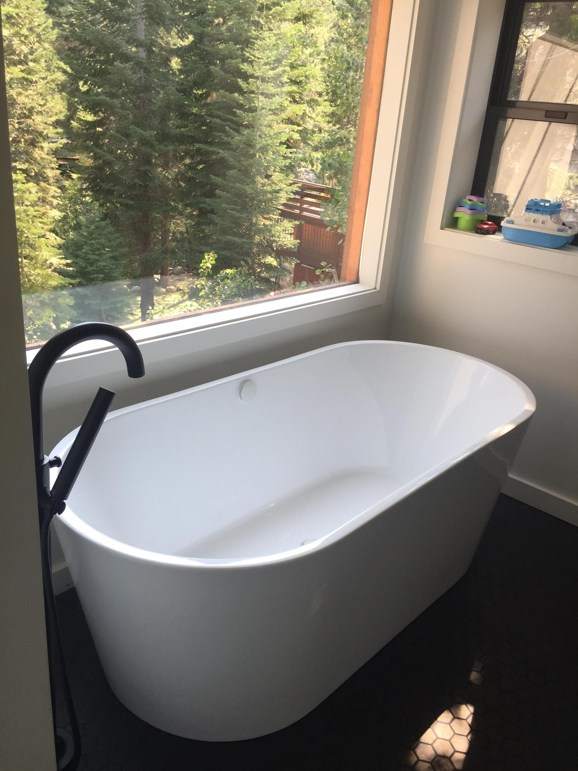 Freestanding white bathtub by window overlooking a forest, black faucet, dark tile floor.