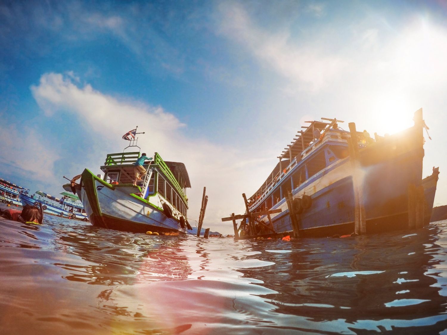 Fishing boats floating on calm water at sunrise, with bright sun glare and a low shoreline in the distance