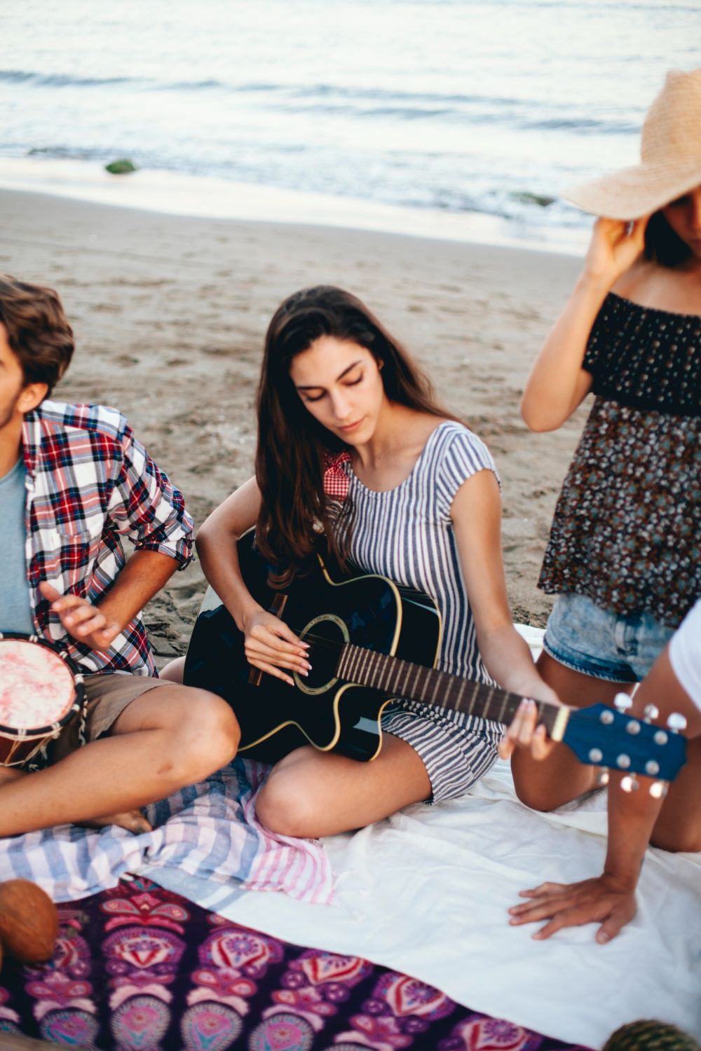 Friends sitting on a beach blanket, playing guitar and drums by the ocean