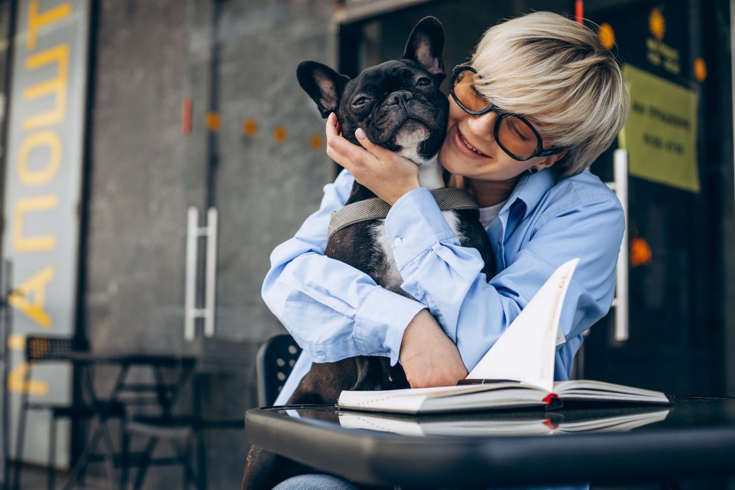 Person hugging a small black dog at a café table with an open notebook, smiling affectionately