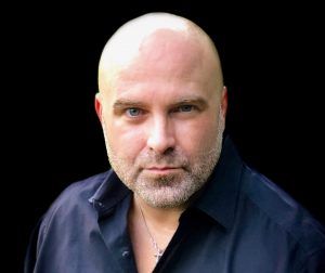 Close-up portrait of a bald man in a dark shirt against a black background.