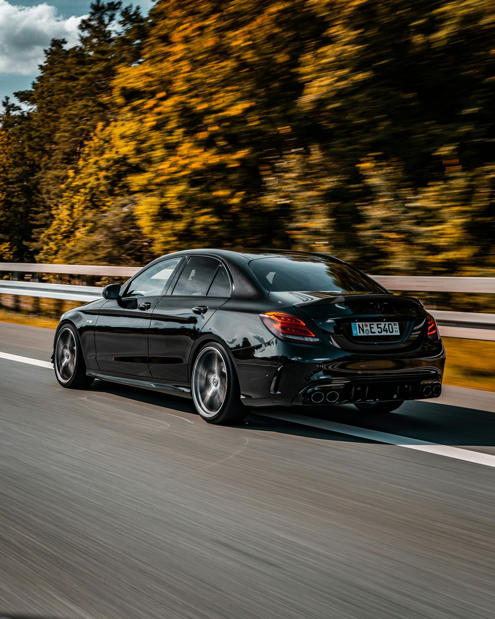 Black sports sedan driving on a highway beside autumn trees and a guardrail