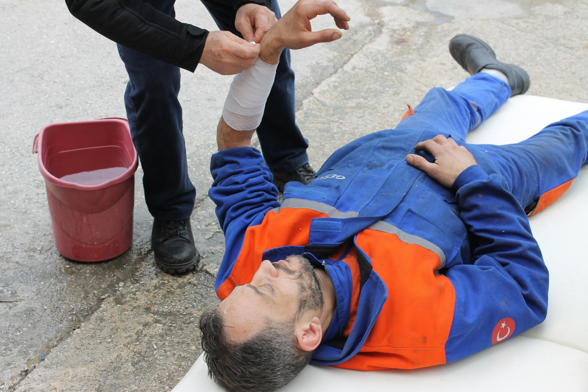 Person in blue-and-orange work clothes lying on the ground while another holds their arm near a red bucket