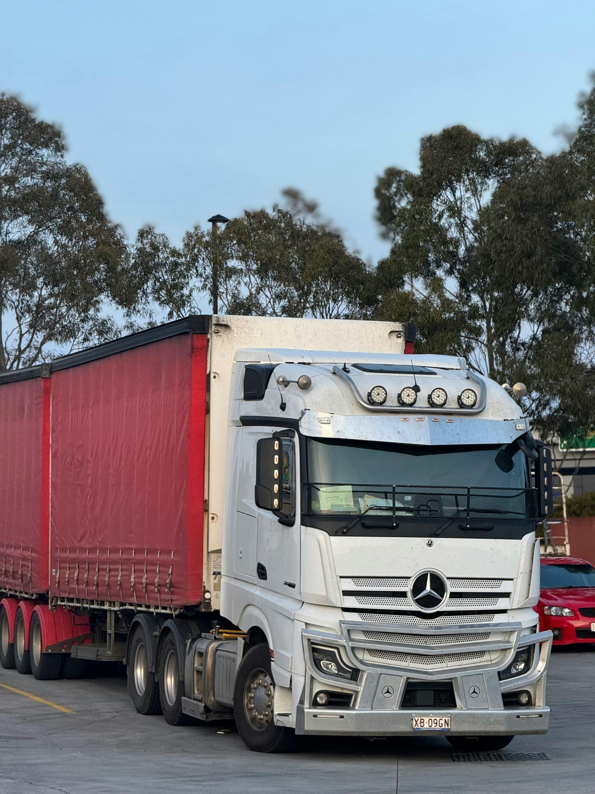 White truck with red trailer on a road, parked outdoors under trees.