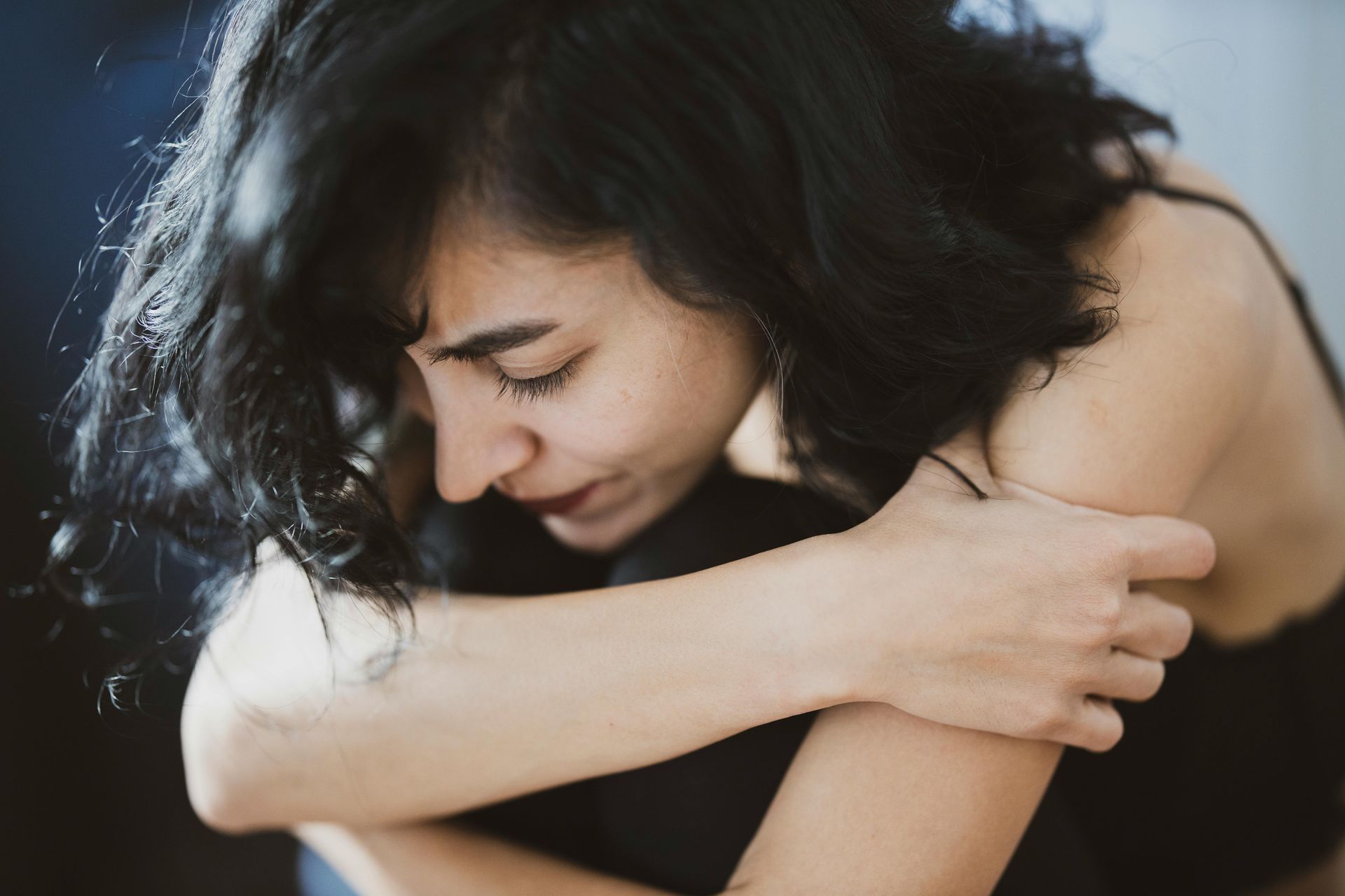 Person with closed eyes hugging their knees, with dark hair and a pained expression, against a blurred background