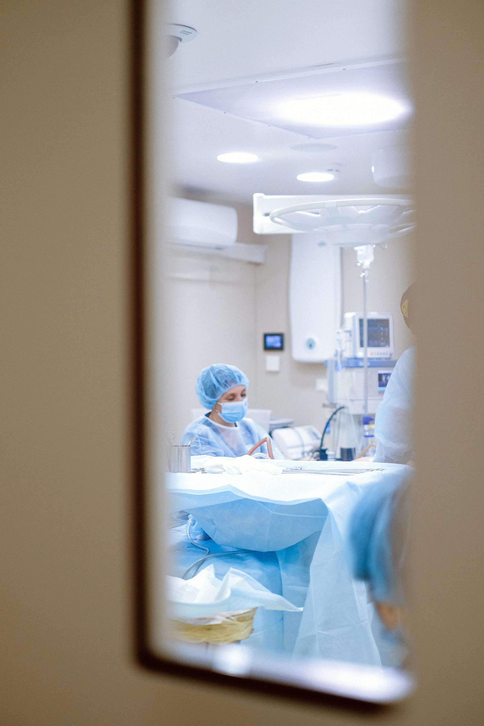 Medical staff in a hospital operating room, seen through a doorway, with blue surgical drapes and bright lights