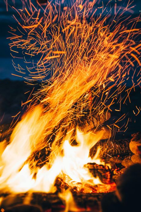 Bright orange flames and sparks rising from a campfire against a dark background