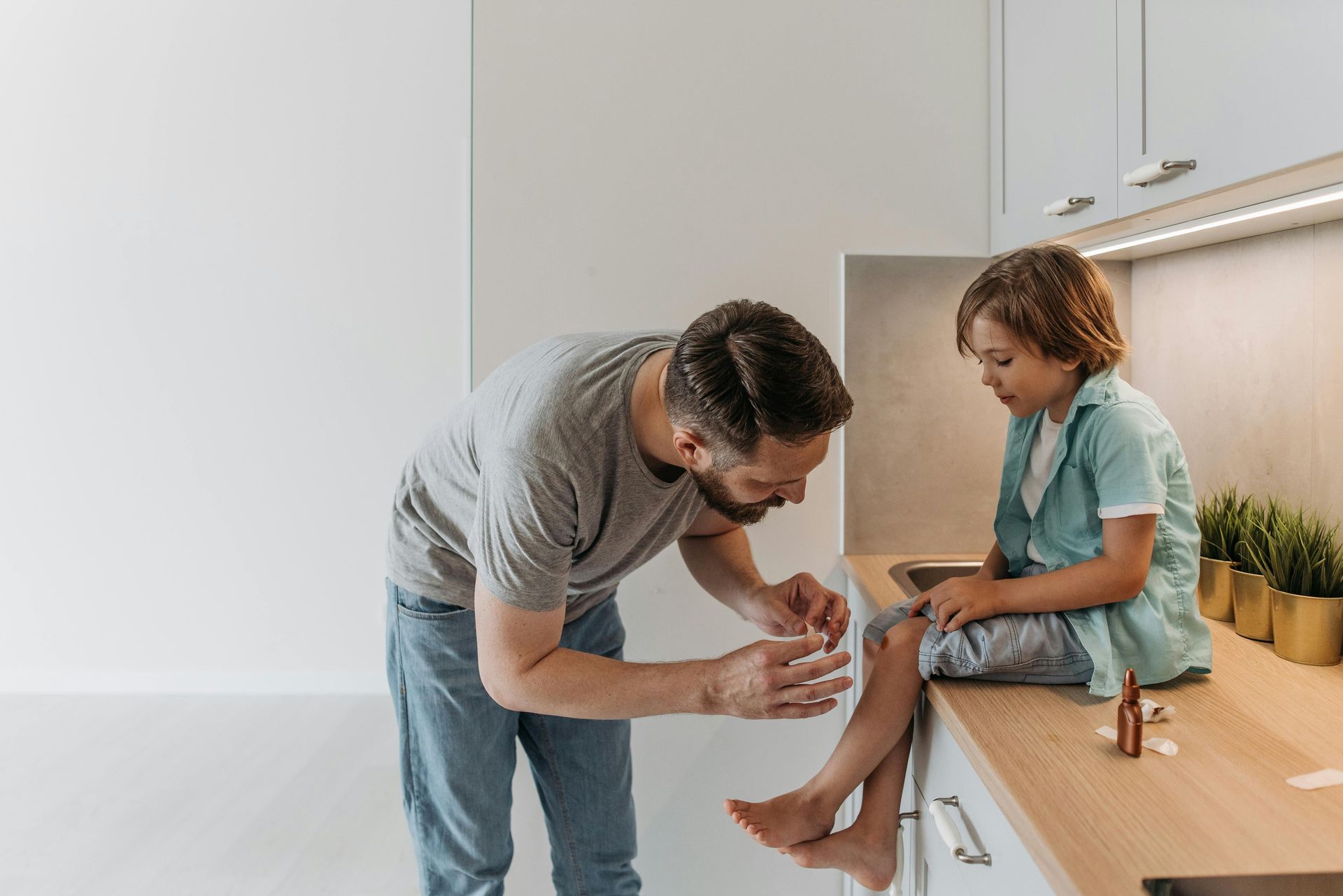Man kneels to bandage a child’s leg on a kitchen counter in a bright room.