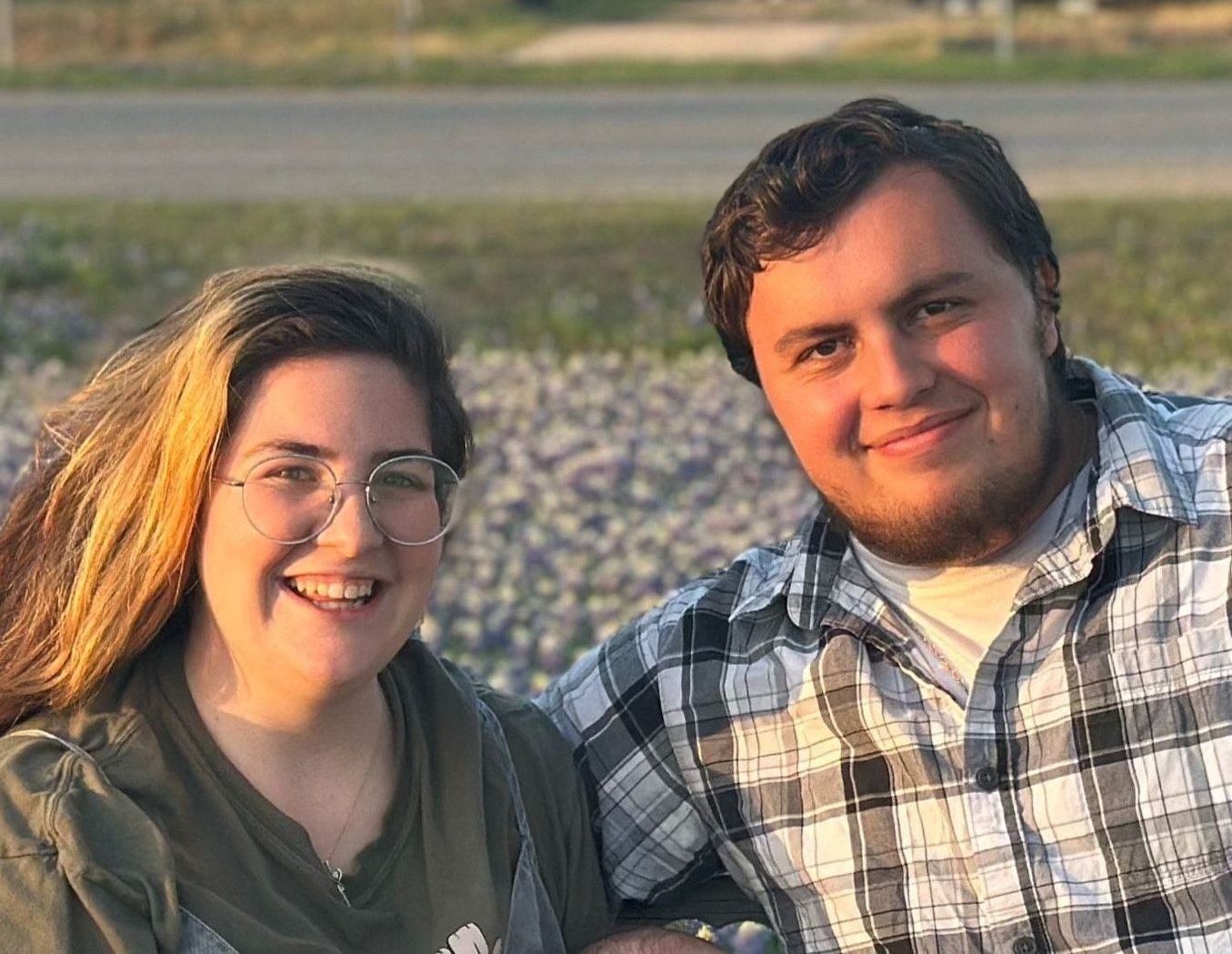 Two people smiling, outdoors. One wears glasses and a green jacket, the other a plaid shirt.