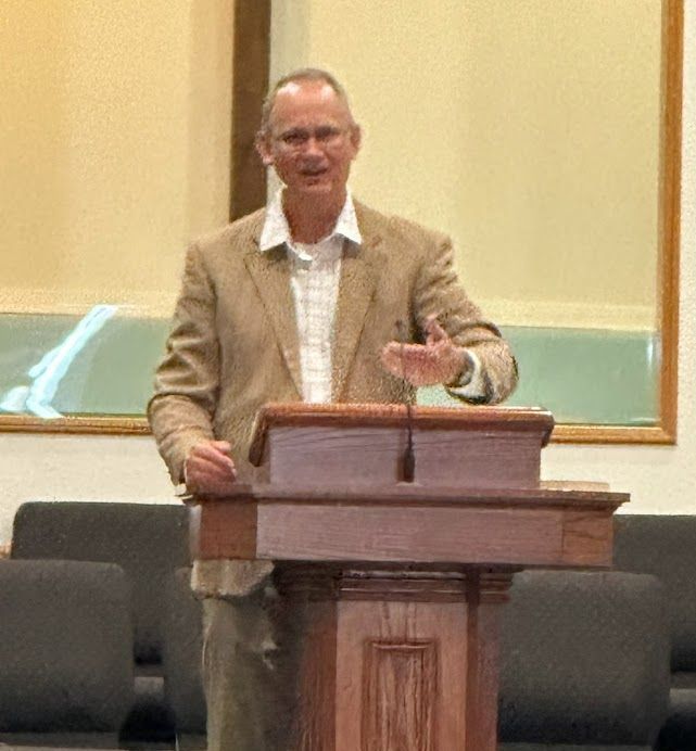 Man in tan jacket at a podium, gesturing. Beige walls, dark podium.