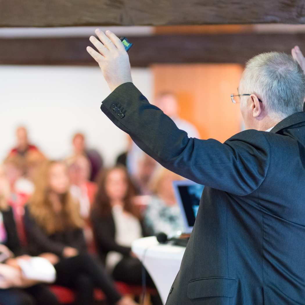 Man giving a presentation, arms raised, to an audience; interior, white walls, and wood beams.