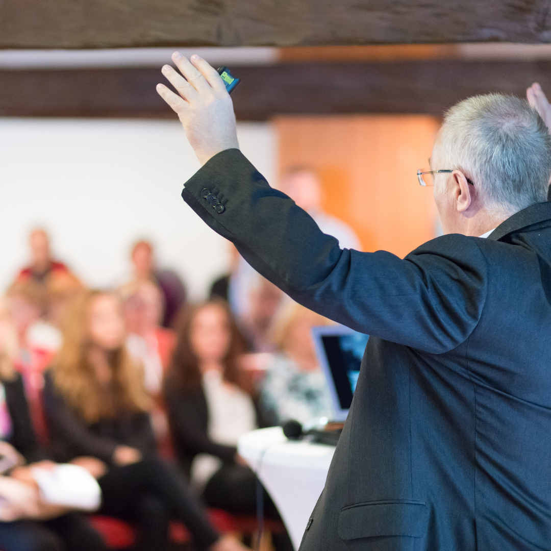 Man giving a presentation, arms raised, to an audience; interior, white walls, and wood beams.