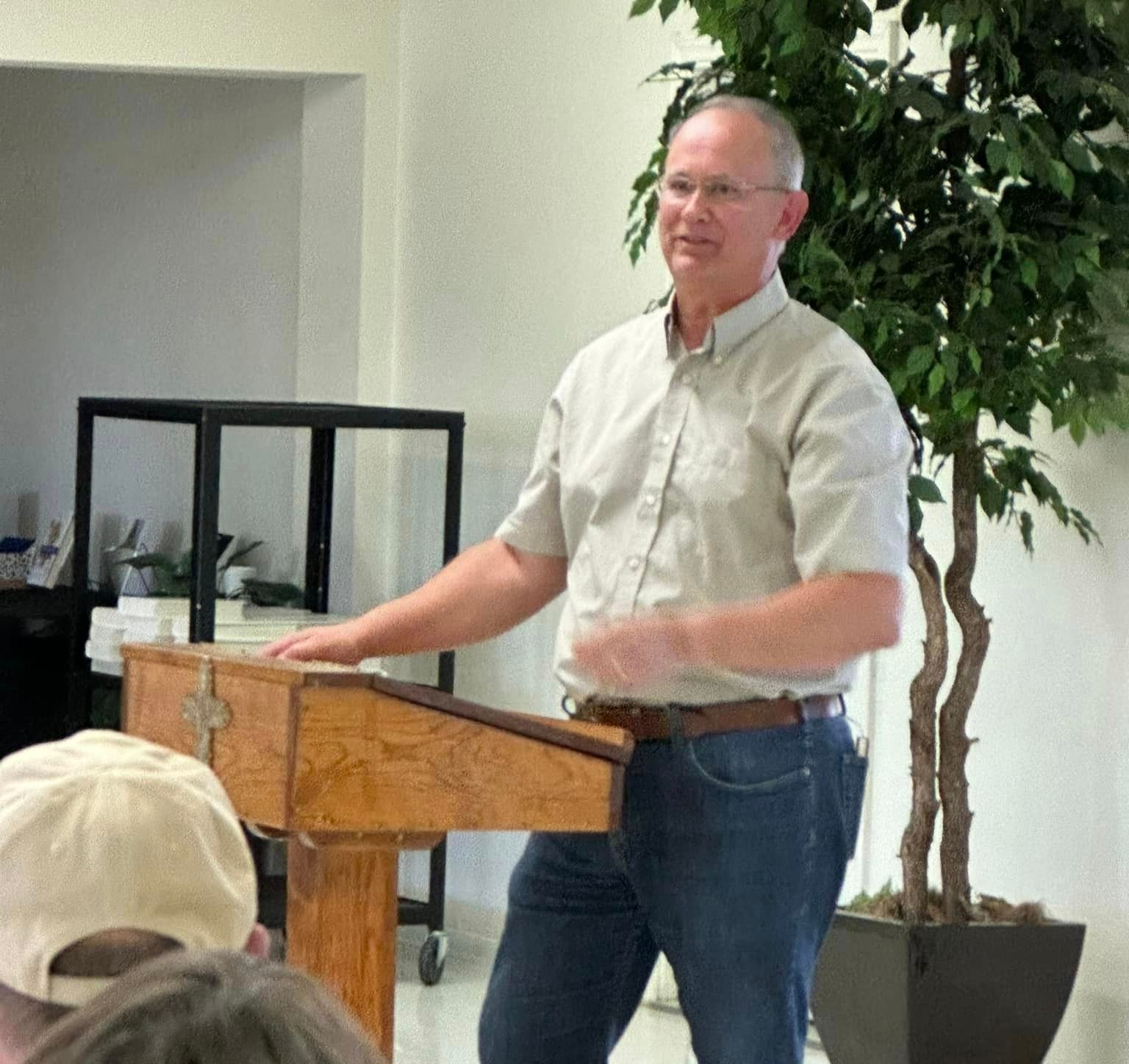 Man speaking at a wooden podium, gesturing with his hand. He wears a light shirt and jeans. A plant is to the right.