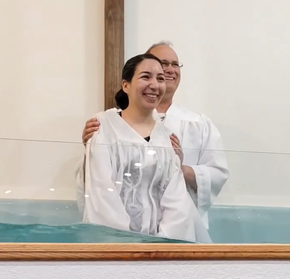 Woman being baptized in a church baptismal pool. A man in white robe stands behind her, smiling.