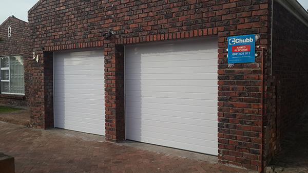 Two white garage doors in a brick building. A blue and white sign is mounted on the right side.