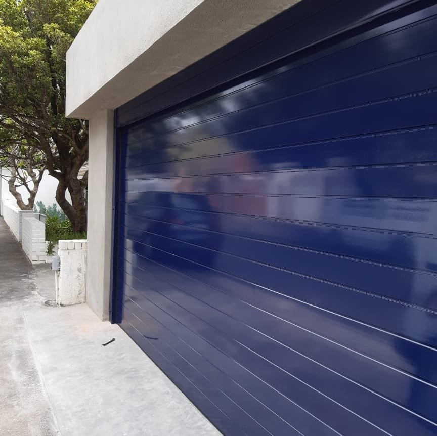 Blue, horizontal-slat garage door. Concrete curb and wall. Road to the left, trees in the background.