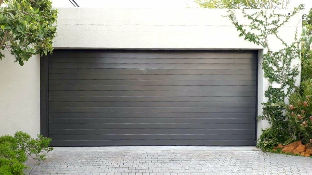 Gray garage door with horizontal lines, flanked by green foliage and a cobblestone driveway.