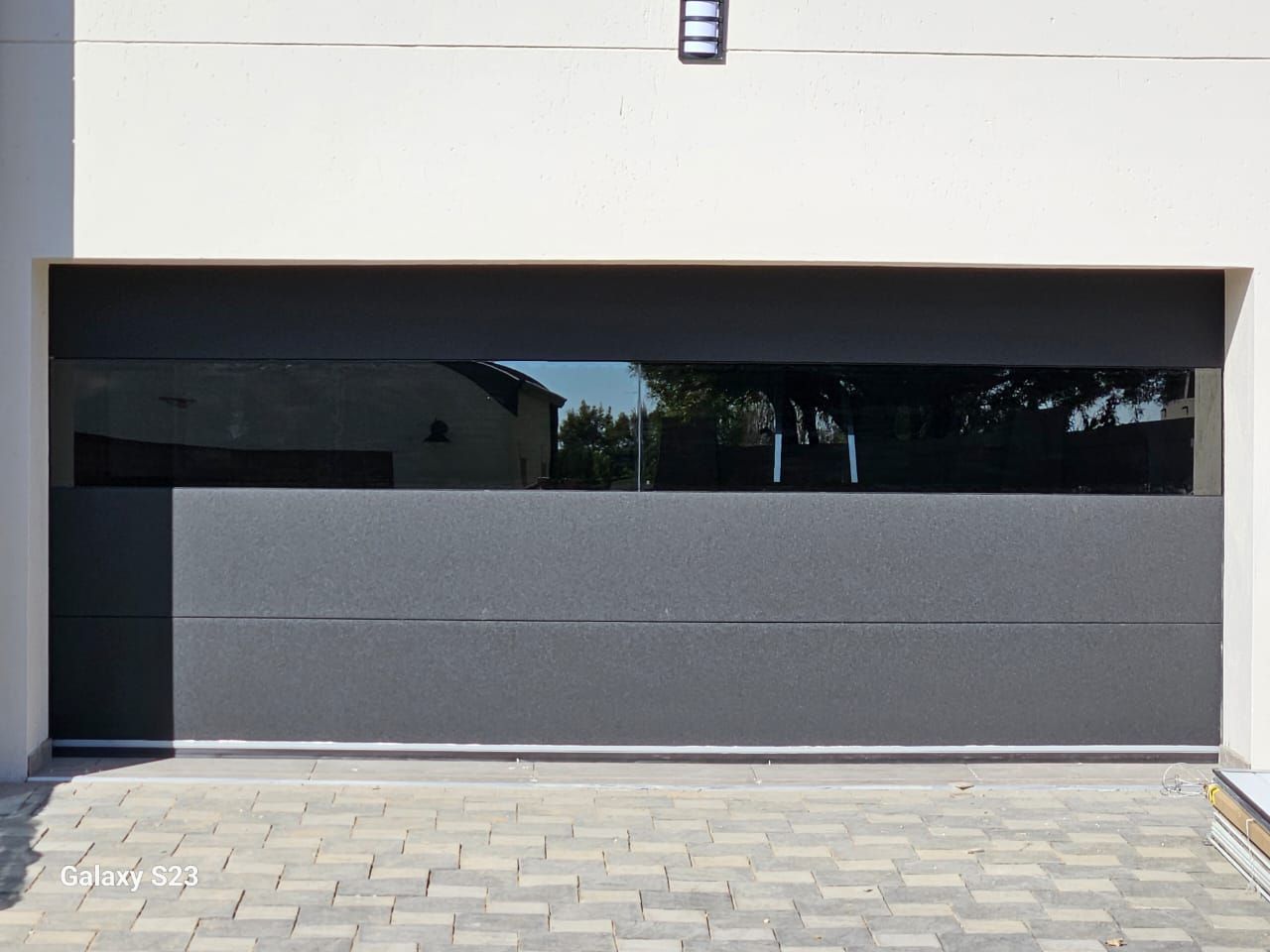 Gray garage door with a horizontal window, set in a white building. Cobblestone driveway.