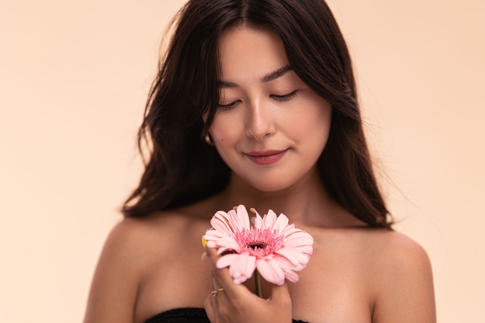 Woman holding pink flower, eyes closed, smiling gently against a peach backdrop.