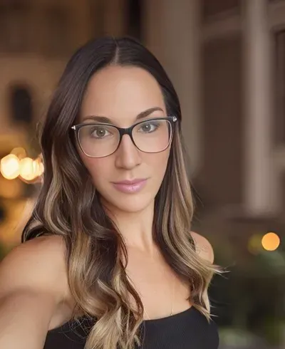Portrait of a person wearing glasses and a black top, standing indoors with a softly blurred background