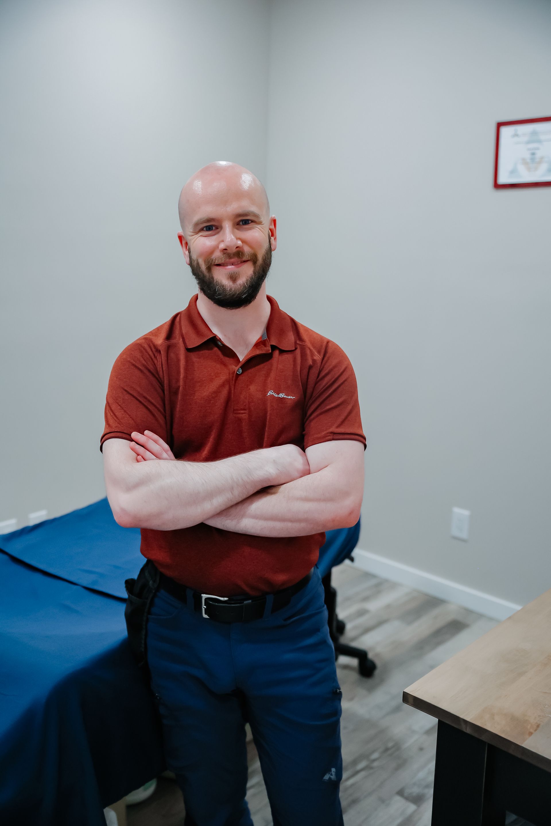 Man standing with arms crossed in a clinic room beside an exam table and desk