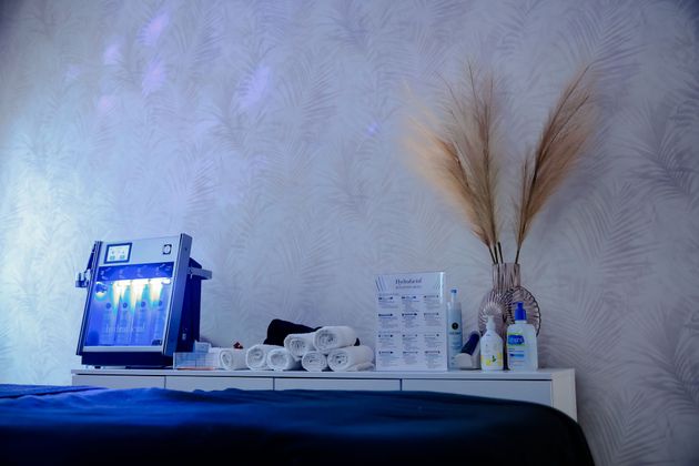 Blue-lit facial spa device on a shelf beside skincare items and dried pampas grass against a textured wall